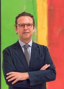 Photograph of Alex Faequharson who is standing in front of a brightly coloured artwork