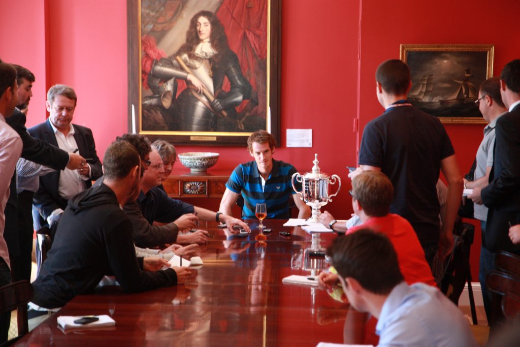 Tennis player Andy Murray seated with his US Open trophy in the dining room of the Consul General in New York surrounded by people. The GAC's painting of the Duke of York hangs on the wall behind him.