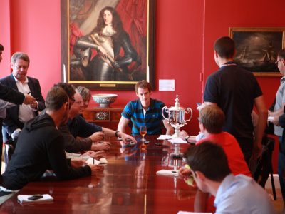 Tennis player Andy Murray seated with his US Open trophy in the dining room of the Consul General in New York surrounded by people. The GAC's painting of the Duke of York hangs on the wall behind him.