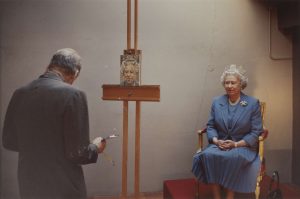 A photo of an artist (Lucian Freud) with his back to the viewer, in front of an easel. An older lady (Queen Elizabeth II) sits in front of the easel, wearing a blue suit.