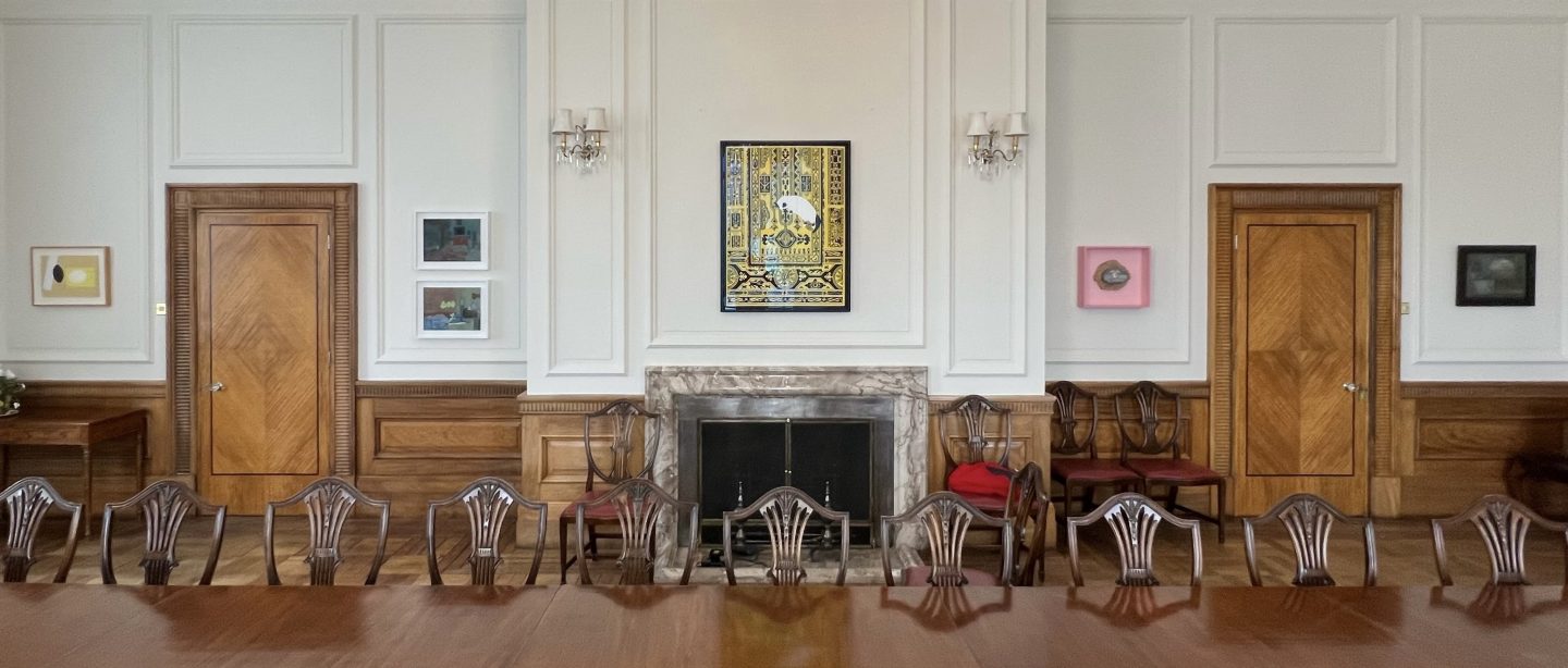 A view of the display in the British Ambassador's Residence in Ankara, Turkey. A polished, wooden dining table, with ten chairs visible; behind it are two wooden doors and works of art hanging on the wall; a vivid black and yellow print in the centre and four smaller ones farming the doors