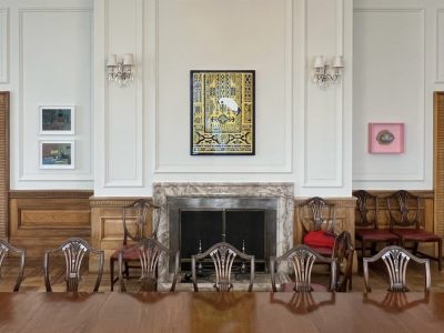 A view of the display in the British Ambassador's Residence in Ankara, Turkey. A polished, wooden dining table, with ten chairs visible; behind it are two wooden doors and works of art hanging on the wall; a vivid black and yellow print in the centre and four smaller ones farming the doors