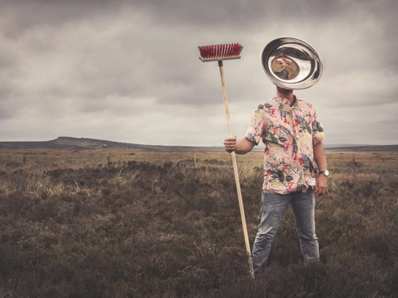 Posing outdoors in the Peak district under a grey sky, the artist wears a bowl as a mask where the fearful image of the Hindu goddess Kali is reflected with her tongue sticking out, and holds a broom.