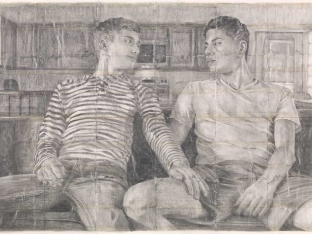 Two men sit on a sofa, with a kitchen behind them, staring intently at each other; the one on the left wears a striped long T-shirt and jeans and his hand rests on the other man's thigh; the second man is wearing a white T-shirt and shorts.