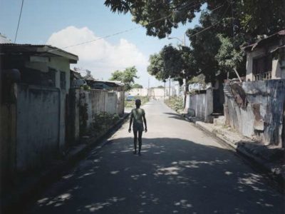 A small child in shorts has its back turned to the camera; the cild is walking down a street, shady under the shadow of a huge tree.