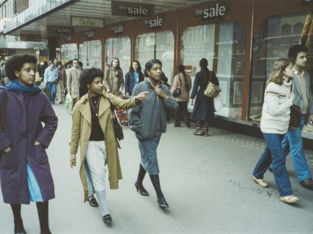 A scene outside a department store; people in coats walk past the shop windows, glancing momentarily inside; those closest to the camera are three black women pointing at something out of shot, and a white man and woman, walking just ahead of them.