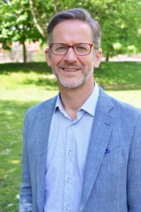 A photo of a white man standing in what looks like a park; he has grey hair and is wearing glasses, a light blue shirt and a blue jacket.