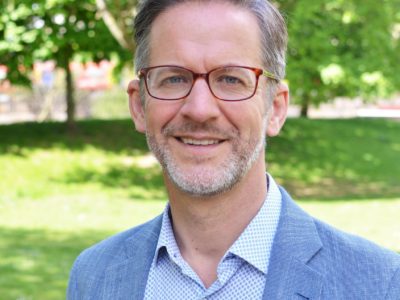 A photo of a white man standing in what looks like a park; he has grey hair and is wearing glasses, a light blue shirt and a blue jacket.