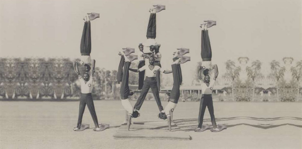 a sepia-toned photograph of twelve men, wearing dark trousers and white T-shirts. They are balanced on each other's bodies – feet on heads, heads on heads – and are seemingly supported by a repeated disembodied hand pointing to the left