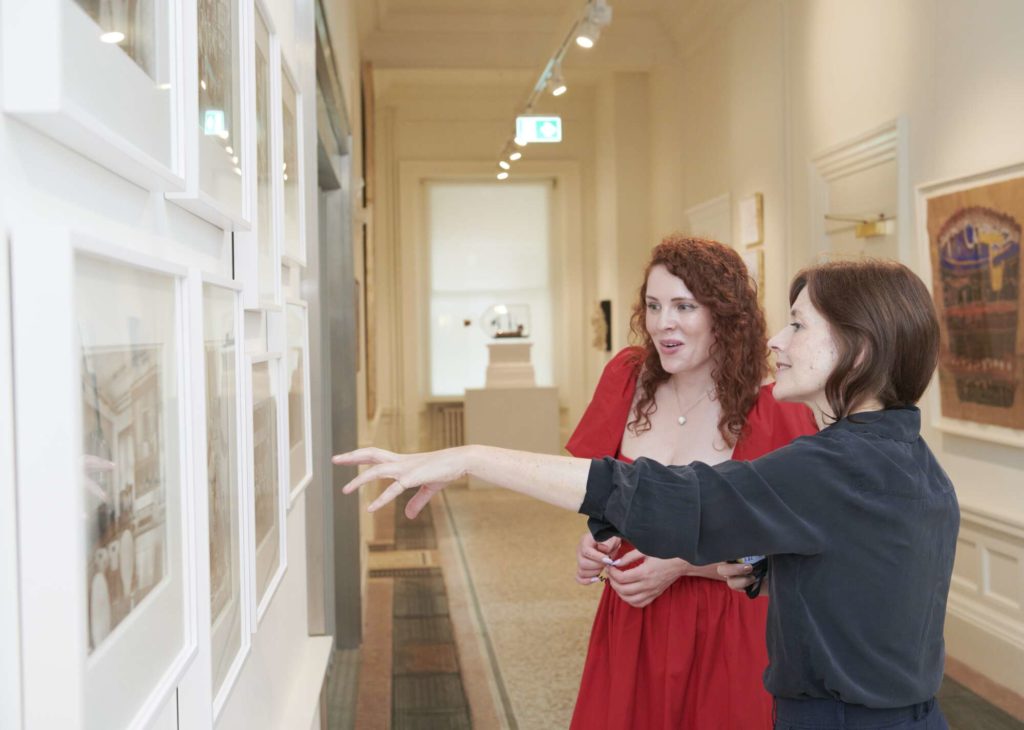 A woman in a red dress with red curly hair stands in front of another woman wearing black. They are in conversation, standing in a corridor surrounded by works of art.