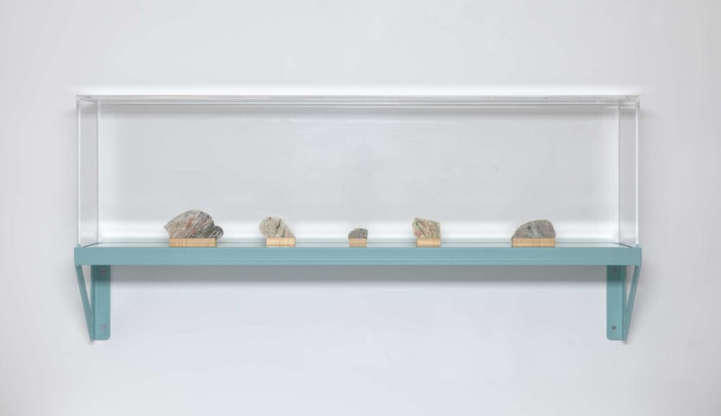 A glass case on top of a blue-green shelf. Inside the case are five stones.