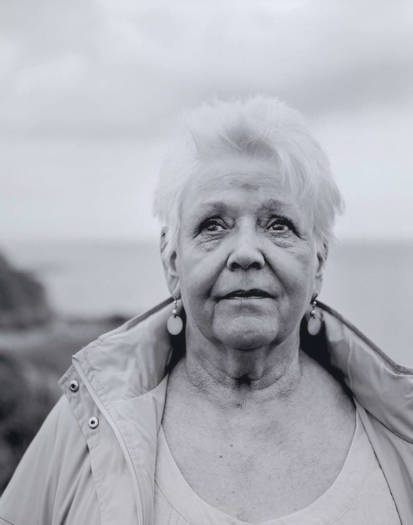 A black and white image of a woman with short white hair, looking just off camera wearing a winter coat in front of the sea