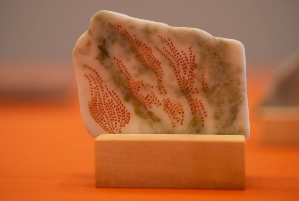 A close-up image of a stone with orange and grey details stood upright, on an orange surface.