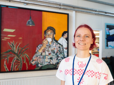 A white woman with pink hair smiling at the camera. Behind her is a painting of a man sipping coffee.